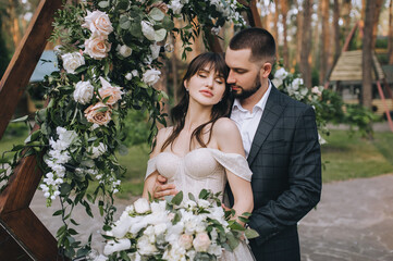 Stylish bearded groom in a suit and a cute brunette bride in a white dress with a long veil tenderly hug on the background of a wooden arch decorated with flowers in nature. Wedding portrait.