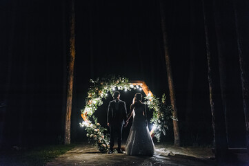 Stylish, young groom in a vest and a beautiful bride in a white long dress stand holding hands near...