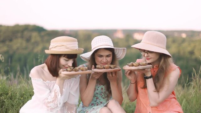 Group of likable female friends in dresses and hats, having picnic in a field on a sunny day, enjoying lunch time, smelling roasted snails on wooden plates.