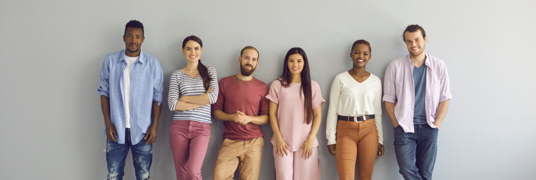 Group portrait of smiling young models in comfortable casual everyday natural color T shirts and jumpers. Happy diverse people posing against light grey studio wall. Banner. Clothes, fashion concept