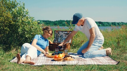 A happy elderly couple is sitting on the grass in a park by the river and drinking wine, celebrating the date on a sunny summer day.