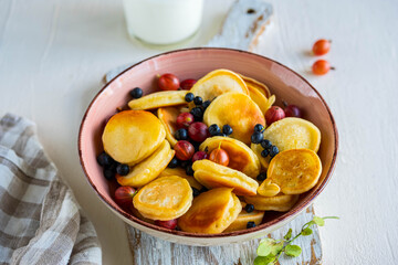 Mini pancakes with gooseberries and blueberries in a pink bowl on a light background. American cuisine.