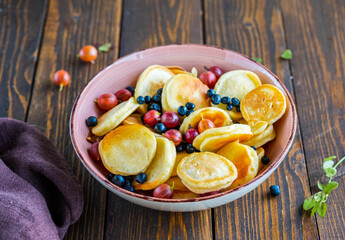 Mini pancakes with gooseberries and blueberries in a pink bowl on a brown wood background. American cuisine.