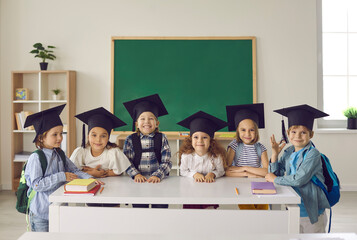 Group portrait of cute school kids. Happy little children in academic caps smiling and looking at camera standing by table with books in new modern classroom - obrazy, fototapety, plakaty
