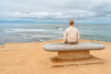 Rear view of a young man sitting on a stone bench by the Pacific Ocean at Sunset Cliffs Nature Park in San Diego, California.