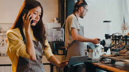 Young Asia female barista taking order by mobile phone and digital tablet standing behind bar counter while talking with customer on mobile phone at cafe restaurant. Owner small business concept.
