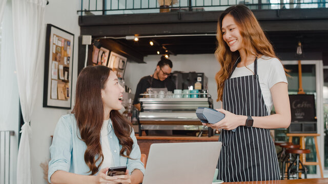 Young Asian freelance women pay contactless at coffee shop. Asian happy girl barista waiter wear gray apron holding credit card reader machine for customer using mobile phone scan pay in cafe.