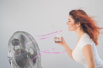 A red-haired Caucasian woman chills by the electric fan and drinks a cold drink. Climate control in...