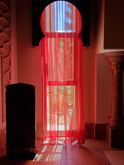 Interior details in retro Arabic style. Sunlight shines through red tulle curtains on orient style window in shape of arch. Light and shadow. Mystique red interior of Turkish palace