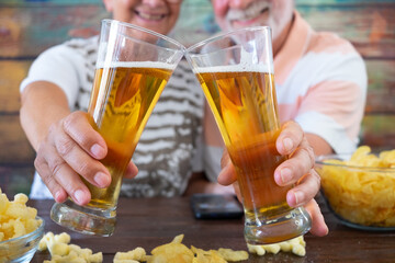 Smiling senior people sitting at pub at a wooden table toasting with two glasses of beer and potato chips.