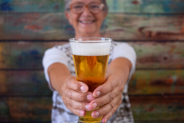 Defocused senior woman sitting at a wooden table holding a glass of blonde beer, looking at camera smiling