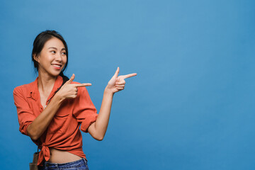 Portrait of young Asian lady smiling with cheerful expression, shows something amazing at blank space in casual clothing and standing isolated over blue background. Facial expression concept.