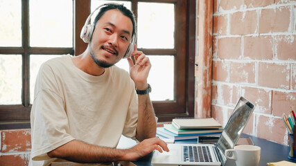 Freelance Asia guy headphones listening music and working on laptop in living room at house in the...
