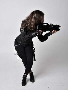 Full length portrait of young woman with natural brown hair,  wearing black leather scifi outfit with corset, standing pose holding a gun on light grey studio background.