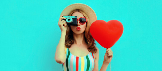 Summer portrait of young woman taking a picture on retro camera with red heart shaped balloon wearing a straw hat on blue background