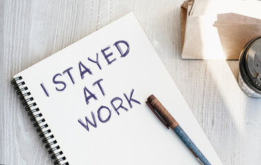 Notepad with the inscription I STAYED AT WORK. Paper cup with coffee and paper bag with breakfast on the table. View from above. Marketing concept.