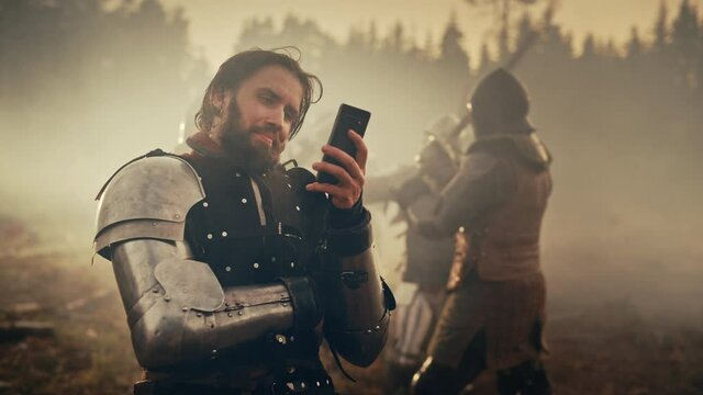 Portrait of Handsome Medieval Knight Using Smartphone on Battlefield, Smiling. Funny Concept: Armored Warrior Having Fun, Ordering Online, Betting, Investing, doing E-commerce. War is Raging