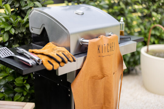 Modern gas grill with leather apron and gloves stands at backyard