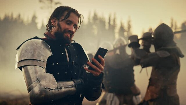Portrait of Handsome Medieval Knight Using Smartphone on Battlefield, Smiling. Fun Concept: Successful Armored Warrior Ordering Online, Betting, Investing, doing E-commerce. War is Raging