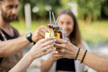 People clinking bottles with a strong alcohol drinks on a picnic outdoors, close-up. Holding bottles of liqueur or tinctures