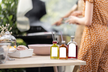 Bottles with liqueur or berry tinctures on a table with food for bbq, people grilling on a background, close-up on bottles with blank labels for copy paste
