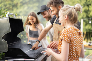 Happy young friends hanging out together, grilling vegetables and meat on a modern grill at picnic. People cooking food outdoors