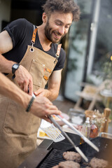 Funny and cheerful man in apron grilling meat and buns for burgers on a gas grill at backyard on...