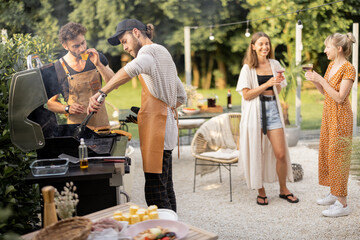 Happy young friends hanging out together, grilling food on a modern grill at beautiful backyard of a country house. People cooking food outdoors