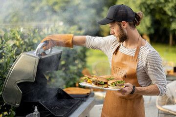 Handsome man in cap and apron making burgers on a grill at backyard. Cooking outdoors and american...