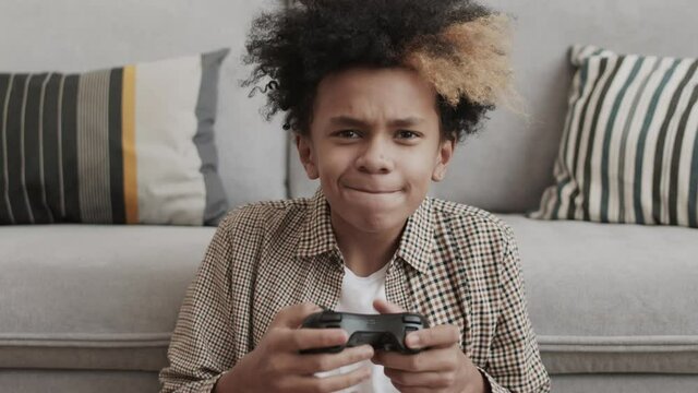 Chest-up of thirteen-year-old Afro boy sitting on floor by couch at home, holding game console controller, playing and getting excited about winning