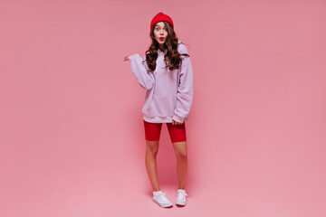 Cool brunette girl in red hat, cycling shorts and purple hoodie whistles. Curly woman with bright lips poses on pink isolated background.