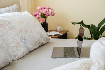 Good morning concept. Empty unmade bed with ranunculus flowers. Close up shot of beautiful spring bouquet and laptop with open lid in bedroom interior. Copy space, background.