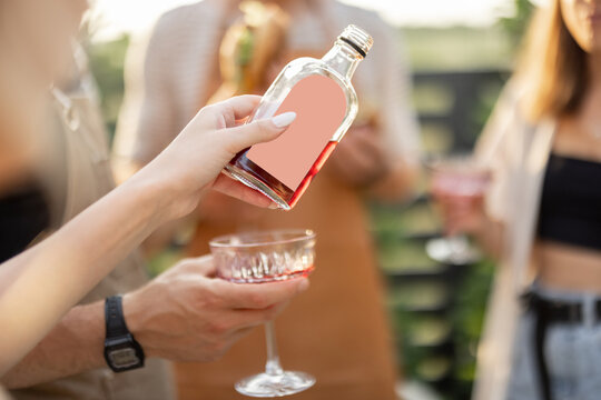 Young people with alcohol drinks at picnic, pouring wine or liqueur into a glasses. Image focused on the bottle with blank label to copy paste