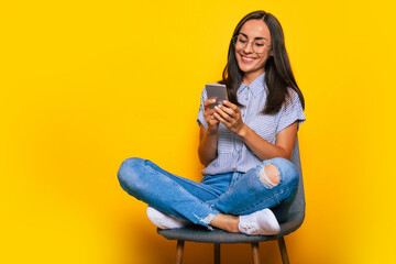 Happy beautiful stylish woman is sitting on the chair and using her smart phone while she is isolated on yellow background