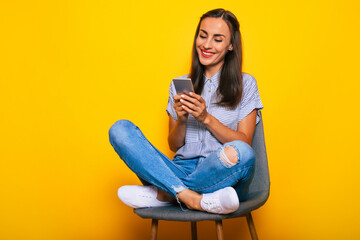 Obraz premium Happy beautiful stylish woman is sitting on the chair and using her smart phone while she is isolated on yellow background
