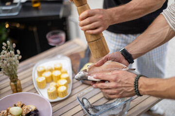 Two men preparing fresh fish for grilling, seasoning it with lemon and spices at table outdoors, close-up