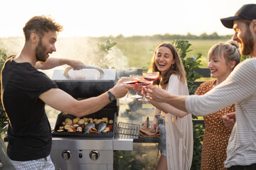 Young friends having fun at picnic outdoors, grilling food and drinking wine at the backyard on a sunset. Spending summer time in a small group on the open air