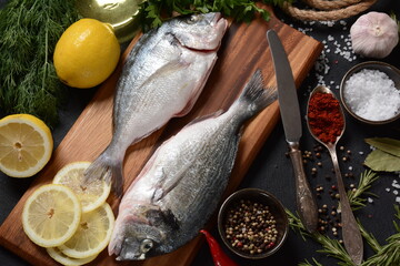Raw fresh uncooked dorado or sea bream fish with lemon, herbs, oil, vegetables and spices on rustic wooden board over black background