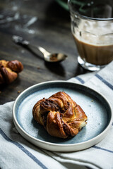 Cinnamon yeast knot bun on blue plate and glass of coffee with milk on dark wooden table.