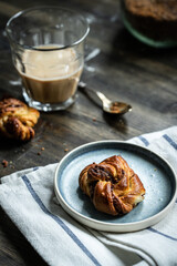 Cinnamon yeast knot bun on blue plate and glass of coffee with milk on dark wooden table.