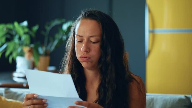Young woman siting on couch holding documents papers feels. Frustrated girl reads financial loss notice, bank debt bill, failed exam test results or subpoena