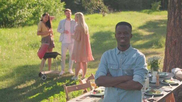 High angle view portrait footage of young African American man standing with arms crossed in park looking at camera, his friends gathering on background