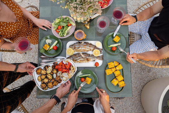 People eating at beautifully served table with grilled vegetables and fish decorateed with field flowers outdoors, top view
