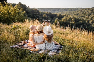 Back view of company of three young female friends having fun, drink red wine, raising glasses and enjoy green hill landscape and beautiful sunset at summer picnic.