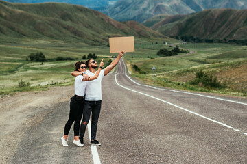 A man and a woman are hitchhiking. Wanderlust, autostop journey concept. Young people traveling hitchhiking. Tourists catches car on road. Hitchhiking couple on the road. Copy space