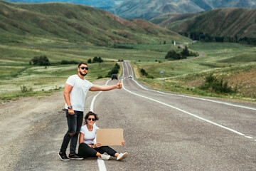 Fototapeta premium Hitchhiking trip. A couple tries to stop a car on the road with an empty cardboard sign, a mock-up. Car travel. Young people traveling hitchhiking. Two young people tourists hitchhiking along a road