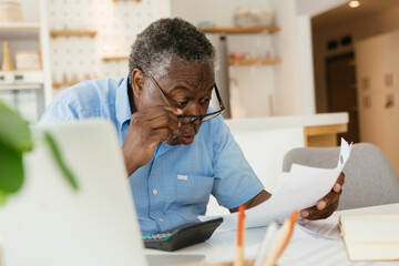 Amazed African pensioner sitting at home and looking at bills he has to pay. He is paying it online...