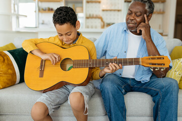 Grandchild sitting with his grandpa in the living room and playing the guitar. Experienced old man...