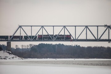a freight train rides on an iron bridge over a frozen river