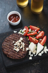 Serbian traditional grilled pljeskavica served with bryndza cheese and red tomatoes, vertical shot on a dark brown stone background